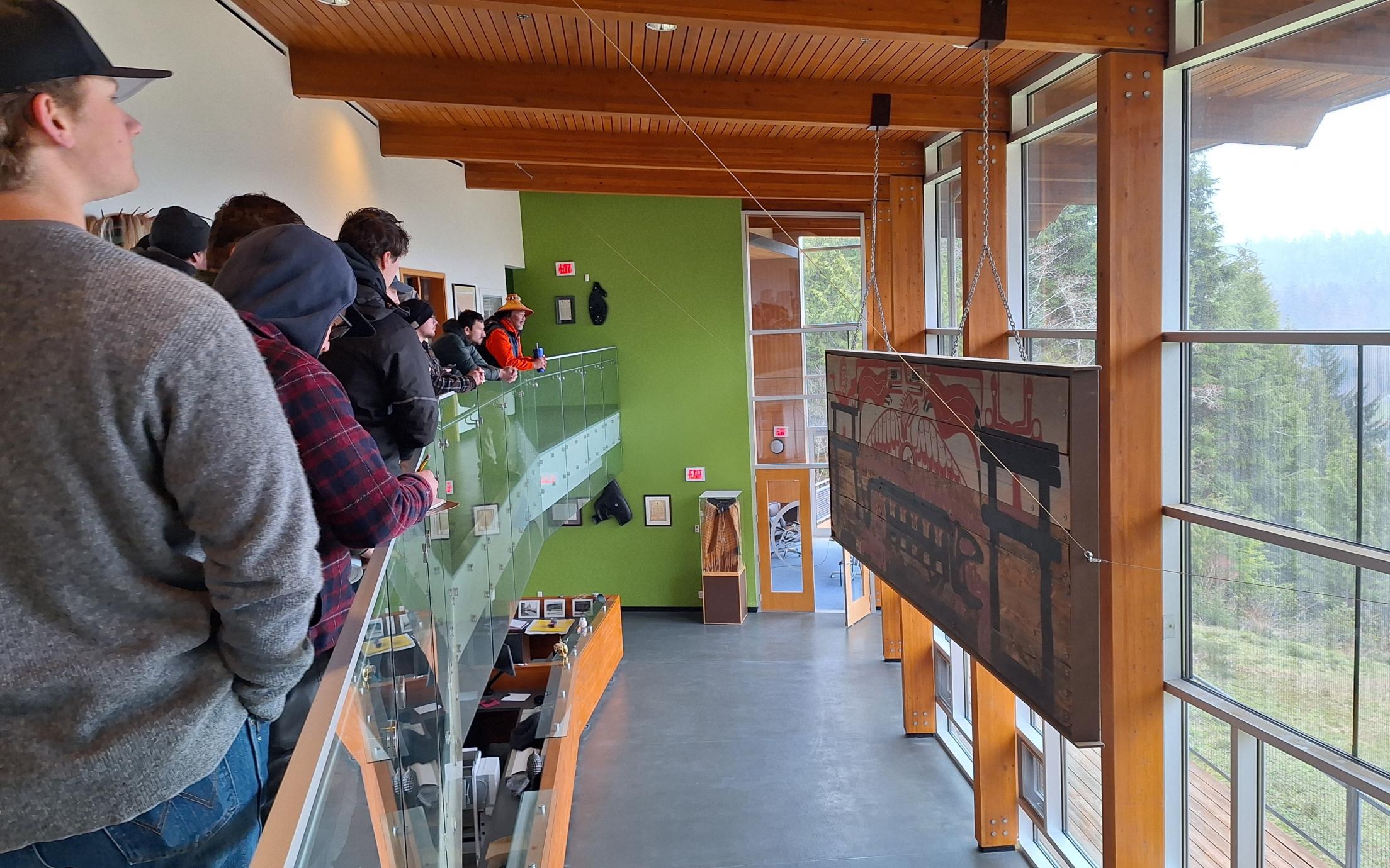 Students look at a piece of art detailing the thunderbird story with the huu-ay-aht nation, in front of a window overlooking a coastal forest.