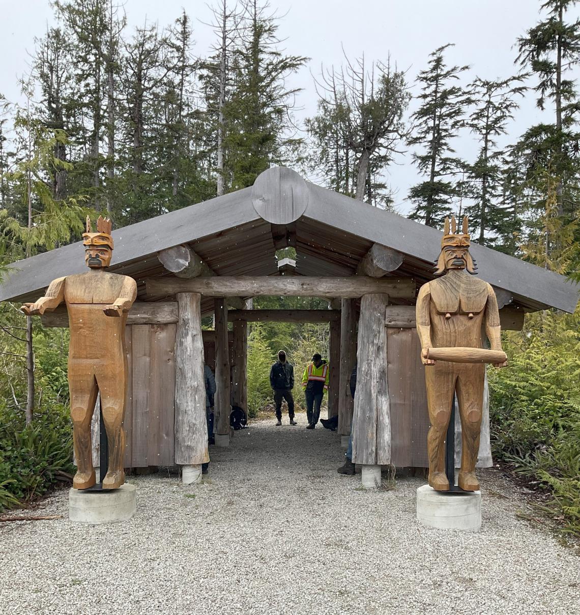 Indigenous longhouse-style building with two wooden statues outside, one of a man with his hands outstretched, one of a woman holding an item. Two students stand in the background through the other side of the building. Trees surround the scene.